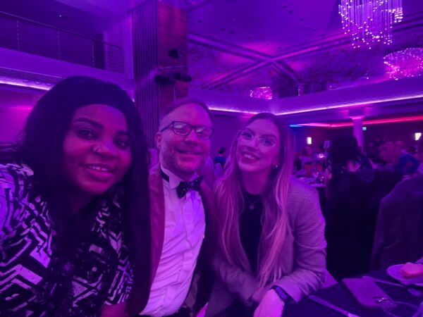 In a pink-hued photo, due to party lighting, Evelyn  - a Black lady with long black hair - is in a selfie with Ross Thomas of Tai Pawb who is a white man with glasses and wearing a blazer and bow tie, and a representative of AICO who is a white woman with long blonde hair and glasses