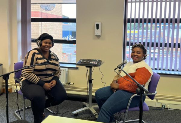 Evelyn - a Black lady in jeans and a jumper - is sat with a microphone in front of her and she is smiling to camera. She is next to a Black lady in a striped jumper who also has a microphone in front of her and is smiling to camera.