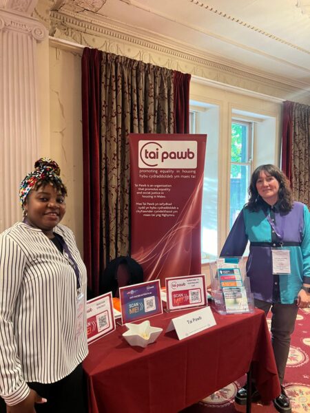 Evelyn - a Black woman in a black and white shirt and a headscarf - and Lynsey - a white woman in a purple and blue shirt with shoulder length brown hair - are stood at a table with Tai Pawb materials and a Tai Pawb banner